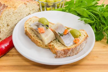 Kahverengi filizlenmiş ekmek, ahşap bir masa üzerinde onun hazırlık closeup için malzemelerin arasında bir daire üzerinde yeşil zeytin ve turşu Atlantik ringa balığı filetosu dilim açık sandviç yaptım