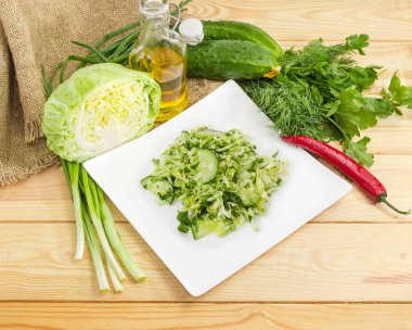Dilimlenmiş taze genç lahana, salatalık ve kare beyaz çanak ve bir açık renkli ahşap yüzeyde hazırlanması için malzemeler doğranmış yeşil soğan sebze yeşil salatası yaptı