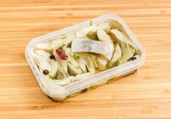 Slices of fillet of pickled Atlantic herring in plastic container with spicy marinade from vegetable oil on a wooden bamboo surface