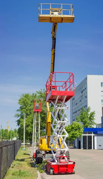 Yellow self propelled articulated boom lift and two red scissor lifts on background of building, trees and sky