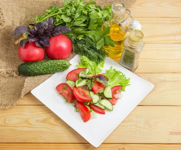 Taze dilimlenmiş domates ve salatalık ve Yeşiller kare beyaz tabak ve ahşap yüzey üzerinde onun hazırlanması için bazı maddeler ile sebze salatası