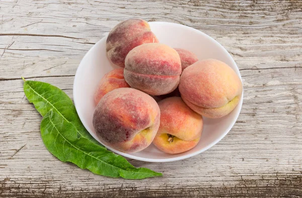 Several ripe fresh peaches in white bowl and leaves of peache tree on an old wooden surface