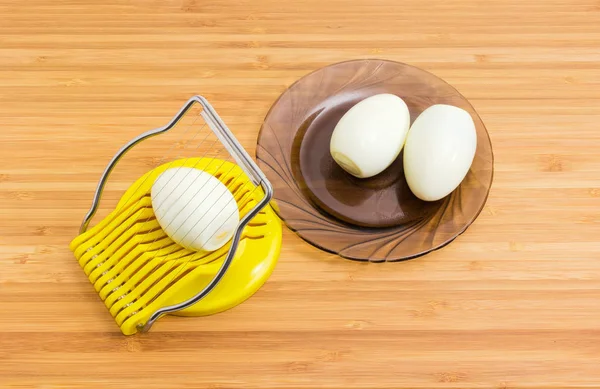 手動エッグスライサーで卵を皮をむいて竹木材表面の暗いガラス受け皿に皮をむいたゆで卵 — ストック写真