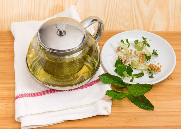 Herbal tea in glass teapot with strainer on napkin and some herbs on saucer and beside on a wooden surface