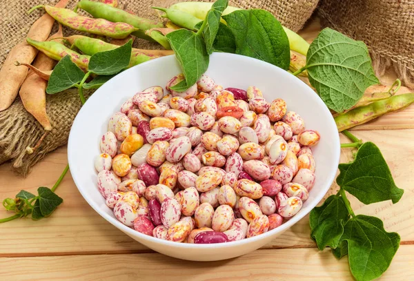 Raw red with white speckled kidney beans in white bowl on a background of bean stems and pods on burlap close-up on a light colored wooden surface