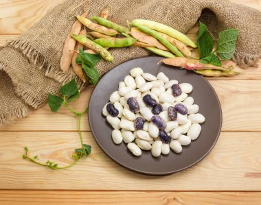 Ham büyük beyaz ve mor benekli böbrek fasulyeli kahverengi tabak, fasulye kaynaklanıyor ve bir çuval bezi ve açık renkli ahşap yüzey üzerinde bakla