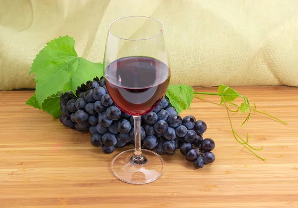 Wine glass of red wine on a background of cluster of blue grapes with vine leaves and tendrils on a bamboo wooden surface