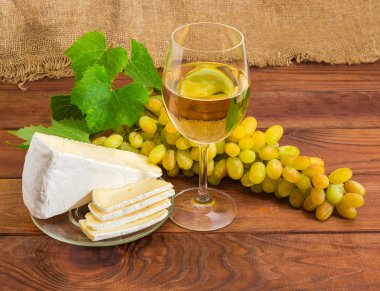 Şarap bardak beyaz şarap ve cam daire beyaz üzüm küme rustik ahşap bir masa üzerinde bir arka plan üzerinde kısmen dilimlenmiş brie peyniri