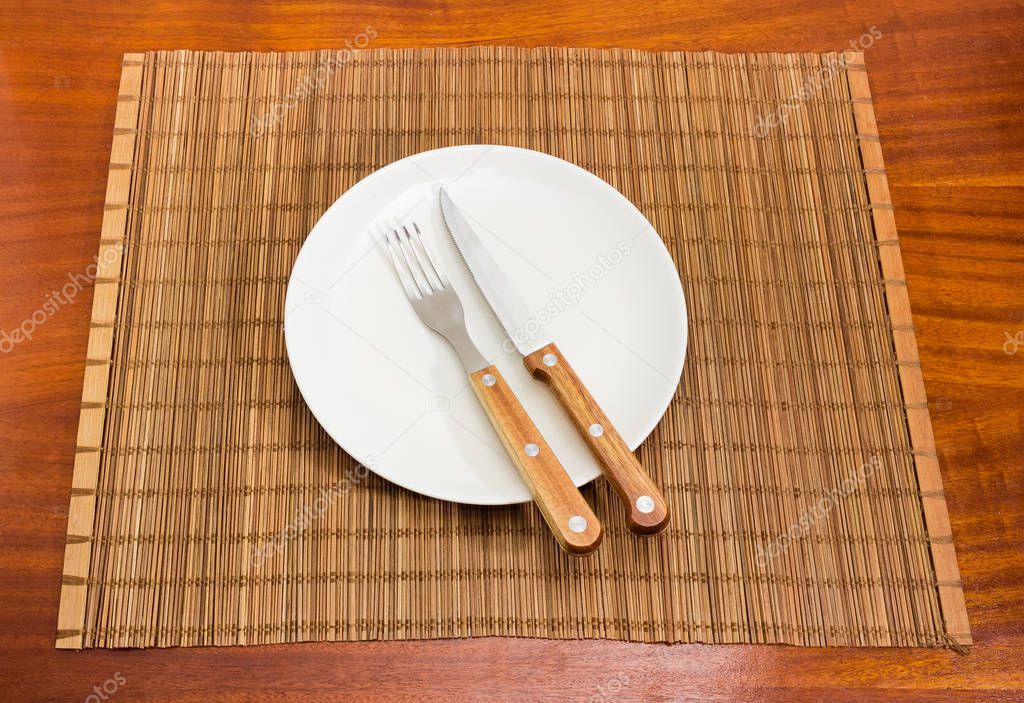 Fork and knife with wooden handles on empty round white dish on the bamboo table mat on a dark red wooden table
