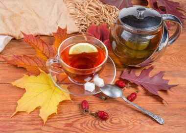 Paslanmaz çelik ve cam Çaydanlık ve bardak fincan sonbahar ile eski rustik ahşap masa kaşık ile uçan daire üzerinde limonlu siyah çay yaprakları, rose hips ve kadın eşarp kurutulmuş