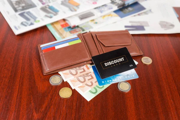 Plastic discount cards lying on open brown leather wallet and money on the red wooden table on a blurred background of goods catalogs