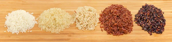 Small piles of raw short-grained white rice, parboiled rice, long-grained brown rice, red rice and black rice on a wooden surface at selective focus
