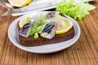 Open sandwich with slices of salted Atlantic mackerel, dill and lemon on brown bread, sprinkled with chopped green onion on saucer on a bamboo table mat close-up at selective focus clipart