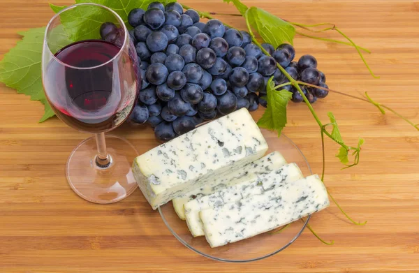 Top view of the glass of red wine on a background of the blue grapes cluster and partly sliced blue cheese on glass saucer on a bamboo wooden surface