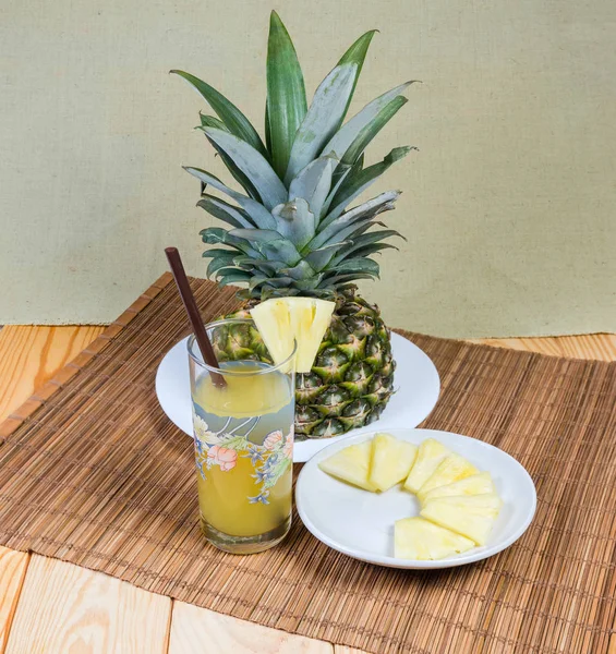 Pineapple juice in glass with drinking straw against of upper part of fresh pineapple with tuft of stiff leaves and pineapple slices on saucer on a bamboo table mat