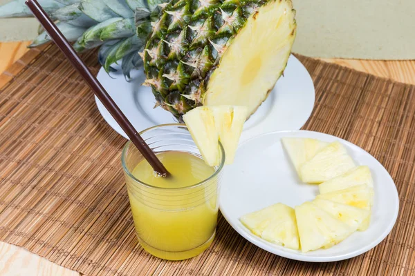 Pineapple juice in glass with drinking straw among of pineapple slices on saucer and upper part of fresh pineapple with leaves on a bamboo table mat