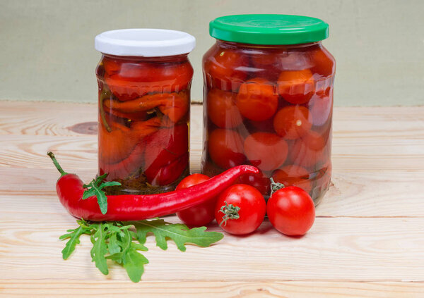 Canned red peppers chili and cherry tomatoes in two different closed glass jars and fresh chili and cherry tomatoes with arugula on a wooden rustic table