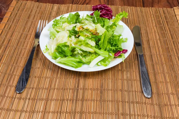 Salad of leaves of green and red lettuce varieties with mustard salad dressing on dish, fork and knife on a bamboo table mat
