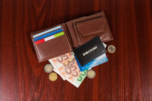 红色木桌上打开的棕色皮革钱包 纸币和硬币上的两张塑料折扣卡的顶视图 — 图库照片