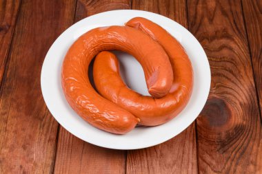 Two ring pork bologna sausages in natural casing on the white dish on dark colored wooden rustic table clipart