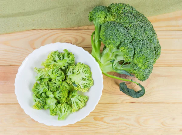Top view of pieces of the fresh broccoli on a white dish and whole broccoli head on light colored wooden rustic table