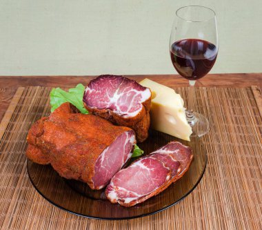 Partly sliced dried pork neck, cheese and glass of red wine on the bamboo table mat on a dark colored wooden rustic table clipart
