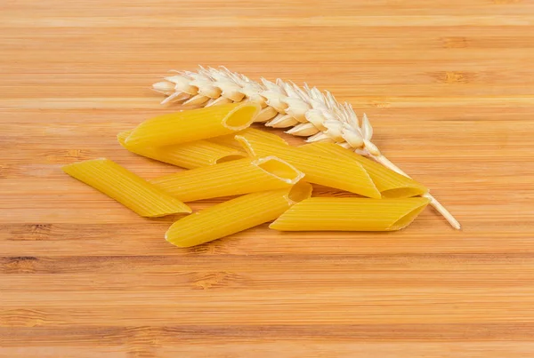 Uncooked penne pasta and wheat ear closeup on wooden surface — Stock Photo, Image