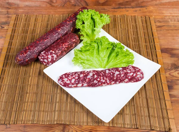 Partly sliced dry-cured sausage on dish on table mat