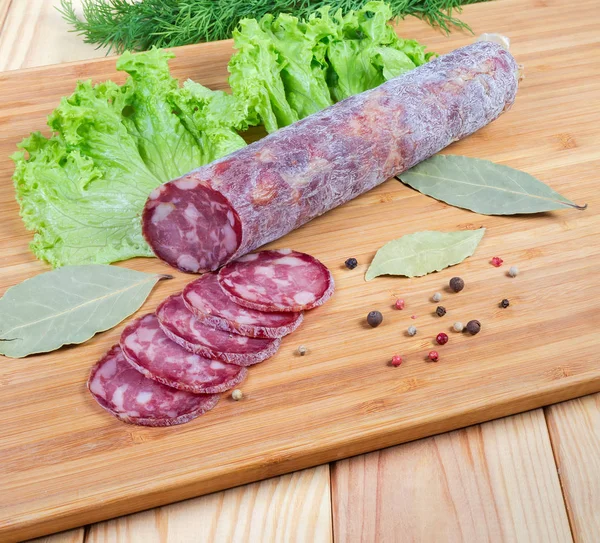 Partly sliced dry-cured sausage, greens, spices on cutting board