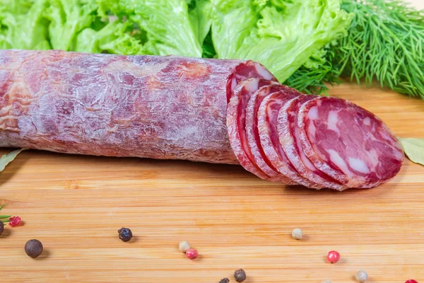 Partly sliced dry-cured sausage among spices against greens