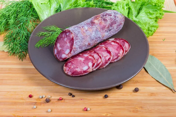 Partly sliced dry-cured sausage on dish among greens, spices