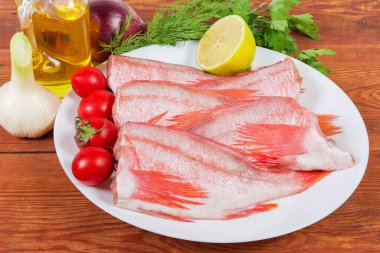 Raw headless gutted carcasses of redfish also known as ocean perch on the dish with lemon and cherry tomatoes, against the vegetables and greens on the rustic table clipart