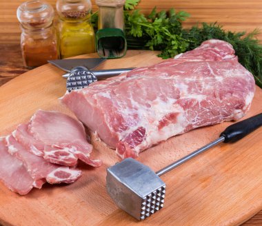 Partly sliced uncooked pork loin, metal meat tenderizer and ax-meat tenderiser on the wooden cutting board clipart