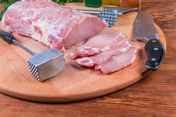 Partly sliced uncooked pork loin among the different meat tenderizers and kitchen knife on the wooden cutting board, close-up in selective focus