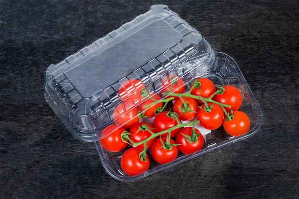 Red cherry tomatoes on branches in the partly open plastic sale packaging in the form of the disposable transparent food container on the black surface