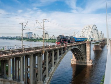 Eski buharlı lokomotifli yolcu treni nehrin karşısındaki kemer köprüsünde hareket ediyor. Darnytskyi Demiryolu Köprüsü Dinyeper Nehri, Kyiv, Ukrayna