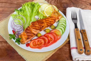 Tabaktaki sebze ve sebzelerin arasında ızgara fileto, kırsal masada çatal ve bıçak.