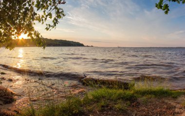 Yaz günbatımı, ağaç dalları boyunca güneş ışınları ve ön planda uzun çimenler ile gölün üzerinde.