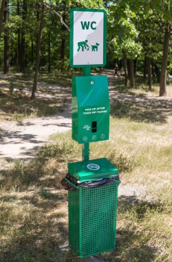 Köpek tuvaleti tabela, poşet dağıtıcısı ve köpek pisliklerini toplamak için çöp kutusundan oluşuyor. Park ağaçlarına karşı.