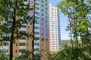 Modern çok katlı apartman kompleksinin cepheleri yazın bir ön plandaki yaprak döken ağaçların gövdeleri ve dalları boyunca görüntülenir.