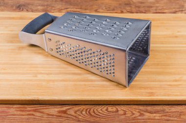 Stainless steel box-shaped four-sided kitchen grater lies on the wooden cutting board with large holes up and very fine grater forward clipart