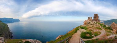 Dik kayalık kıyıları ve eski bir kalenin kalıntıları, uzak bir pelerin ve su yüzeyi bulutlu sabah gökyüzüne karşı, panoramik üst manzara