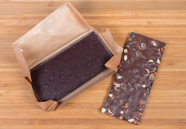 Bar of dark chocolate in open paper wrapper and bar of chocolate with various nuts on the wooden surface, top view