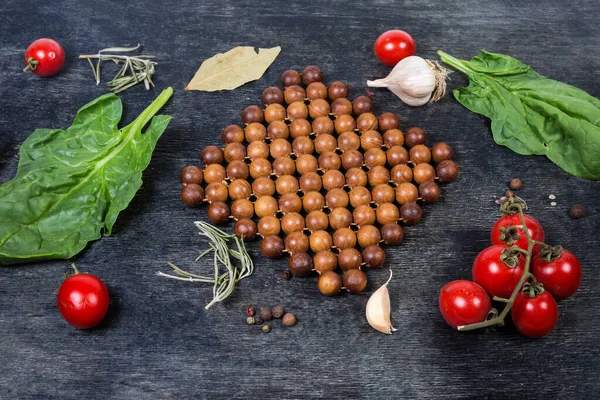 Wooden stand under the hot kitchen utensils made of juniper beads connected into the mat on a dark surface among the spices, tomatoes and spinach