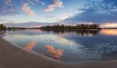 Önplanda kumlu kıyıları olan geniş bir nehir ve karşı kıyıda güneş doğmadan önce sonbaharda ormanla kaplanmış panoramik manzara.