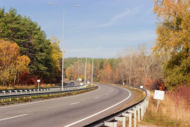 Otoyolun kıvrımlı, yol yüzeyi işaretli, sokak lambaları, trafik bariyerleri ve ormanın her iki tarafında güneşli bir sonbahar sabahı