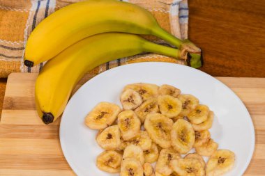 Kurumuş muz cipsleri beyaz tabakta kesme tahtasının üstünde, ahşap bir masada soyulmamış taze muzların bulanık arka planında.