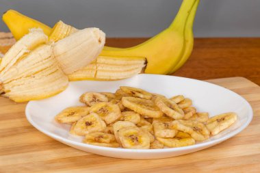 Beyaz tabakta kurutulmuş muz cipsi, kesme tahtasının üzerinde. Kısmen soyulmuş olanının arka planında ve ahşap bir masada taze muzlar.