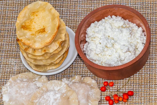 A food flat lay of the fried yellow tortillas with cottage cheese lie like a pyramid on a small white plate and a clay brown bowl with cottage cheese stands on a wicker napkin and a group of not fried tortillas with white flour is near by and a brush