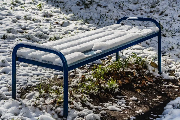 一个明亮的湿木漆成蓝色美丽的长凳与蓝色的金属腿与白雪站在绿草与黄色的叶子和白雪在公园 — 图库照片